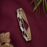 A gold-plated bangle bracelet with alternating rectangular links featuring clear crystals and black inlay, displayed on a textured dark red fabric background with decorative plant leaves.