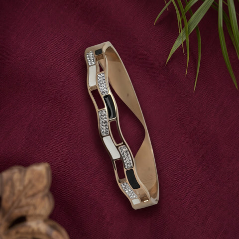 A gold-plated bangle bracelet with alternating rectangular links featuring clear crystals and black inlay, displayed on a textured dark red fabric background with decorative plant leaves.