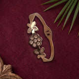 A metallic gold-toned bracelet featuring a grapevine design with leaves and a butterfly motif, displayed on a textured dark red fabric background with decorative green leaves.