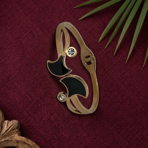 A gold-toned crescent moon-shaped brooch with black enamel sections and two small clear rhinestone accents, displayed on a textured dark red fabric background with decorative green leaves and a wooden object at the edges.
