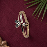 A rose gold bangle bracelet with black enamel details, a black clover motif, and a central clear crystal, displayed on a textured dark red fabric background with green leaves.
