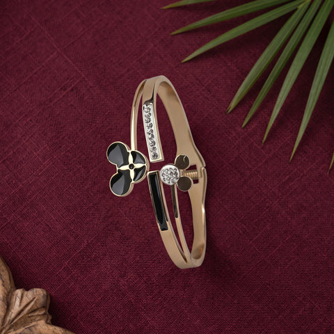 A rose gold bangle bracelet with black enamel details, a black clover motif, and a central clear crystal, displayed on a textured dark red fabric background with green leaves.