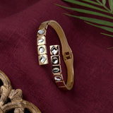 A gold-plated open cuff bangle bracelet with clear faceted gemstones, displayed on a textured dark red fabric background with decorative props.