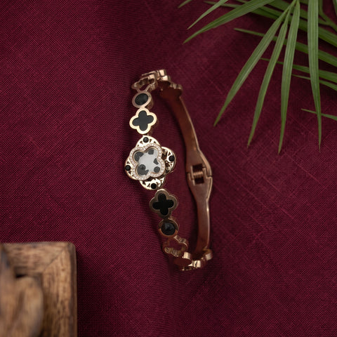 A gold-toned bangle bracelet with clover motifs, featuring black inlay on three clovers and a central white clover with black detailing, displayed on a textured dark red fabric background with a green plant leaf in the corner.