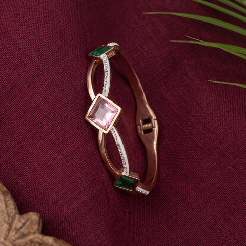 A rose gold bangle bracelet with a central light pink square gemstone, accented by green emerald-cut stones and a white inlay, displayed on a textured dark burgundy fabric background.