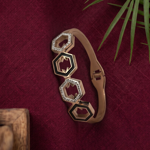 A gold-toned bangle bracelet with three interlocking hexagonal links, two of which are set with clear crystals and one is open to reveal a red interior. The bracelet is displayed on a textured dark red fabric background with a green plant leaf in the corner.