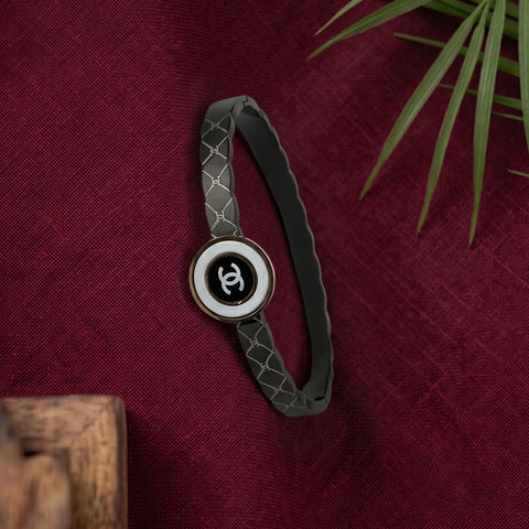 A dark gray bracelet with a woven pattern and a central round charm featuring a black and white interlocking CC logo, styled on a textured burgundy fabric background with a green plant leaf in the corner.