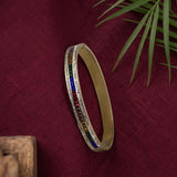 A gold-plated bangle bracelet with a row of colorful gemstones set along the outer band, displayed on a textured dark red fabric background with green palm leaves.