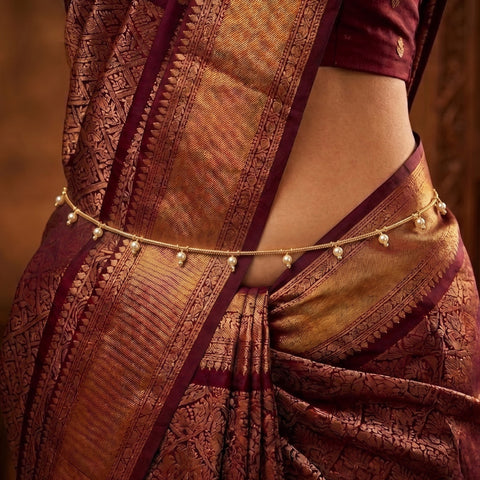 A close-up of a person wearing a traditional sari with rich gold and maroon brocade patterns, accessorized with a delicate gold chain waist belt decorated with small pearl-like beads.