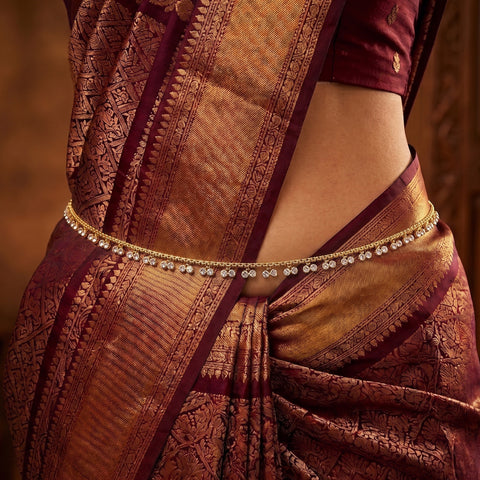 A close-up of a traditional Indian woman wearing a gold-toned chain waist belt adorned with small clear stones, paired with a richly patterned maroon and gold saree and a matching maroon blouse.