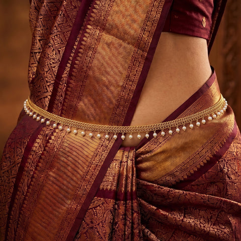 A close-up of a woman wearing a traditional Indian saree with a gold chain waist belt adorned with small white pearls, paired with a deep maroon blouse.
