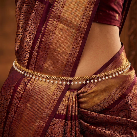 A close-up of a person wearing a traditional Indian saree with a gold-toned chain waist belt decorated with small white pearls, paired with a deep maroon and gold patterned saree and a matching maroon blouse.