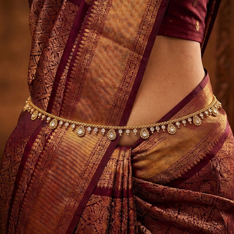 Close-up of a person wearing a richly detailed Indian sari with a gold-toned waist chain adorned with small stones, featuring deep maroon and golden brocade patterns.