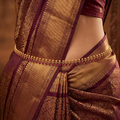 A close-up of a model wearing a traditional Indian saree in deep maroon and gold, accessorized with a gold chain waistband embellished with small red stones.