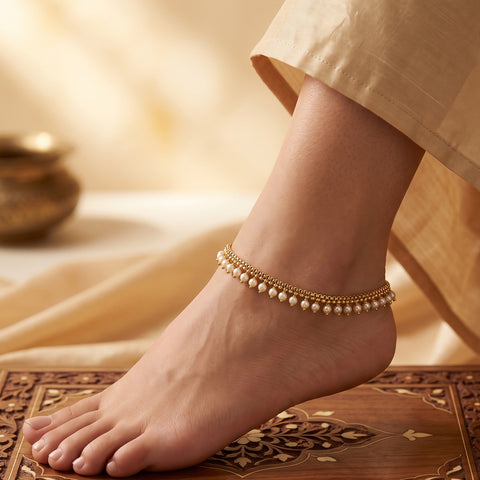 A delicate gold ankle chain adorned with small white pearls worn on a person's ankle, shown in a warm, styled setting with a wooden patterned surface and soft beige fabric.