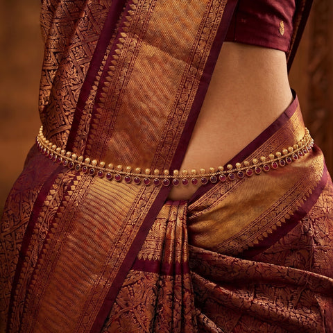 A close-up of a person wearing a traditional Indian saree with a gold-toned waist chain decorated with small red stones, paired with a deep maroon blouse.