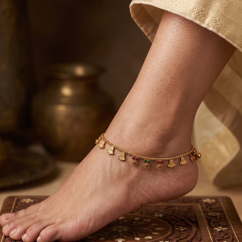 A close-up of a person's foot and ankle wearing a delicate gold chain anklet adorned with small bell-shaped charms and tiny red and green stone accents, standing on a patterned rug with a warm, rustic background.
