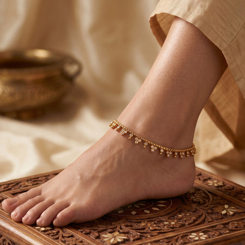 A delicate gold chain anklet with small dangling floral charms is worn on a bare foot resting on an ornate carved wooden surface, with soft beige fabric draped in the background.