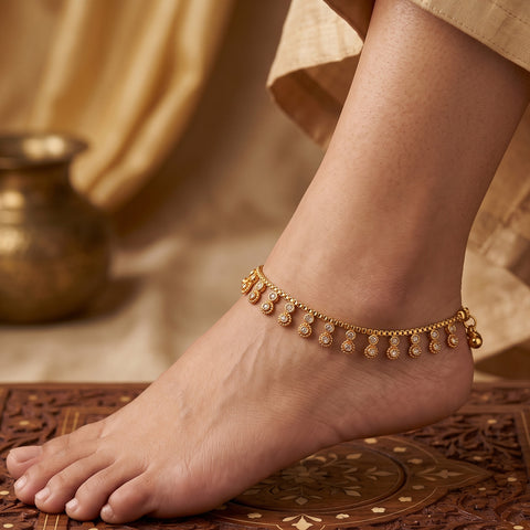 A delicate gold-toned anklet with small dangling bead charms worn on a person's ankle, shown against a warm neutral background with a carved wooden surface and draped fabric.
