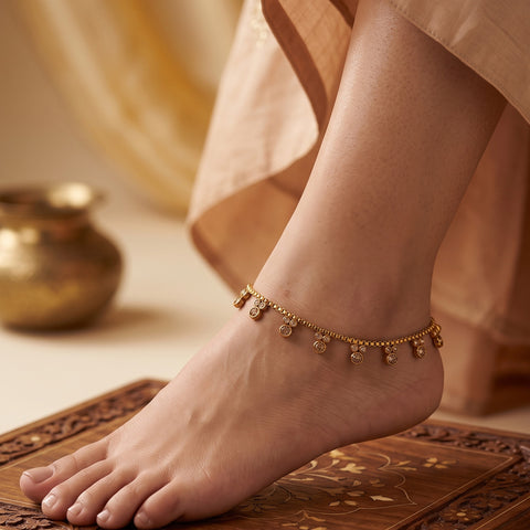 A delicate gold ankle chain bracelet with small hanging charms worn on a person's ankle, shown in a warm, styled setting with a carved wooden surface and soft beige fabric.