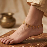 A delicate gold ankle chain with small dangling disc and floral charms is shown worn on a person's ankle, with the foot resting on an ornate wooden surface and a soft neutral background.
