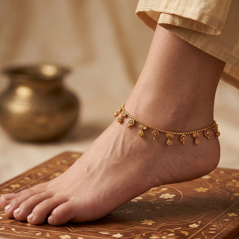 A delicate gold ankle chain with small dangling disc and floral charms is shown worn on a person's ankle, with the foot resting on an ornate wooden surface and a soft neutral background.