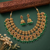 An ornate gold-toned traditional necklace set with matching earrings, decorated with green beads, white pearl-like accents, and red gemstone details, arranged on a dark green background with a carved wooden prop and small dried flowers.