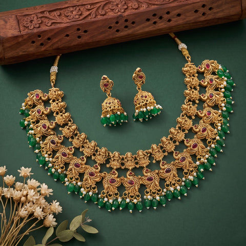 An ornate gold-toned traditional necklace set with matching earrings, decorated with green beads, white pearl-like accents, and red gemstone details, arranged on a dark green background with a carved wooden prop and small dried flowers.