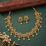 Gold-toned traditional jewelry set with a necklace, matching earrings, and pearl-like drops, decorated with green and purple stones and arranged on a dark green background with a carved wooden prop and dried flowers.