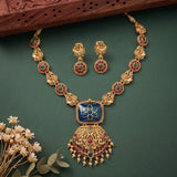 Gold-toned traditional Indian jewelry set featuring a necklace with a blue enamel heart pendant, matching earrings, and ornate gold-toned chain links decorated with red and green stones, arranged on a dark green background with dried flowers and a carved wooden prop.
