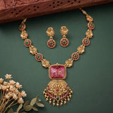 Gold-toned traditional Indian jewelry set featuring a necklace and matching earrings with a large pink rectangular pendant, red and green stone accents, and ornate filigree details, arranged on a dark green background with small dried flowers and a carved wooden prop.