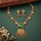Gold-plated Indian traditional jewelry set featuring a necklace and matching earrings with green enamel panels, red stone accents, and gold filigree details, displayed on a dark green background with decorative dried flowers and a wooden prop.