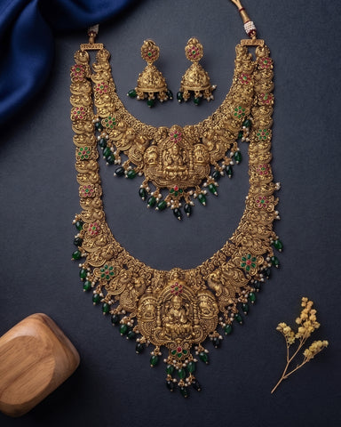 An ornate gold-toned traditional necklace set with matching earrings, decorated with green and red stone accents and hanging bead drops, arranged on a dark navy background with decorative fabric and a small wooden block.