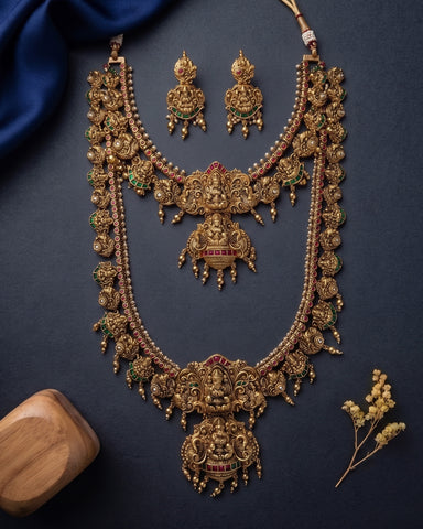 An ornate gold-toned traditional necklace set with matching earrings, decorated with intricate filigree, red and green enamel accents, and dangling bead details, arranged on a dark navy background with decorative fabric and dried flowers.