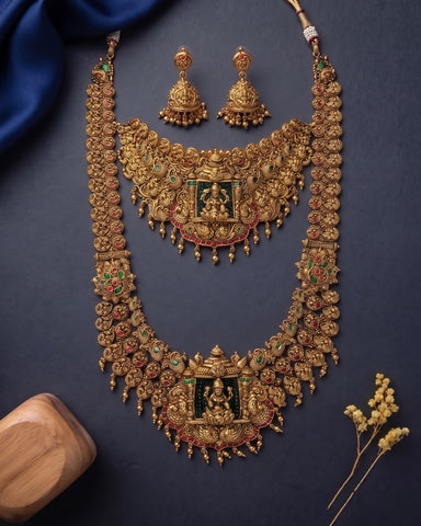 An ornate gold-toned traditional necklace set with matching earrings, decorated with green and red enamel details and small hanging motifs, arranged on a dark blue background with decorative fabric and dried flowers.