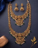 An ornate gold-toned traditional necklace set with matching earrings, decorated with green and pink stones and white pearl-like beads, arranged on a dark navy background with decorative fabric and a wooden prop.