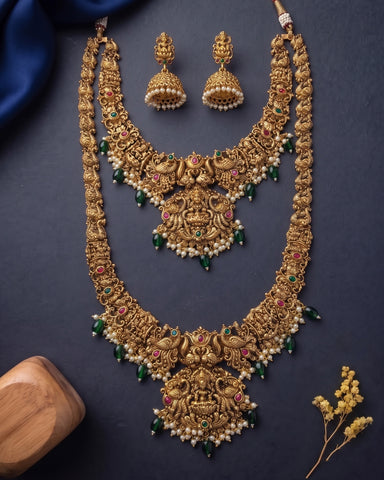 An ornate gold-toned traditional necklace set with matching earrings, decorated with green and pink stones and white pearl-like beads, arranged on a dark navy background with decorative fabric and a wooden prop.