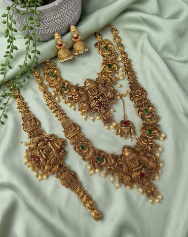 An ornate gold-toned traditional jewelry set laid out on pale mint green fabric, including a necklace, bracelet, earrings, and a matching headpiece, decorated with gold filigree, pearl-like beads, and small green and red stone accents.