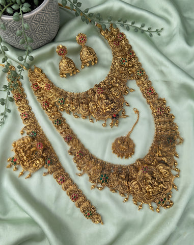 An ornate gold-toned traditional jewelry set arranged on pale mint-green satin fabric, including a large necklace, matching earrings, a bracelet, a pendant, and a small chain, with decorative greenery and a small pot in the corner.