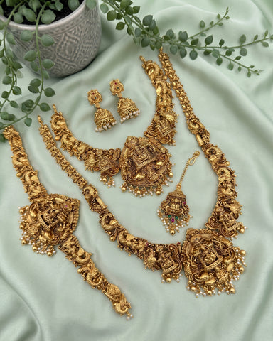 An ornate gold-toned traditional jewelry set arranged on pale mint-green satin fabric, including a necklace, bracelet, earrings, and a pendant, with decorative green leaves and a small pot in the background.