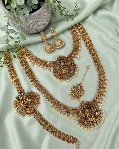 An ornate gold-toned jewelry set arranged on pale mint-green satin fabric, including a necklace, bracelet, earrings, and a pendant, with decorative greenery and a small pot in the corner.