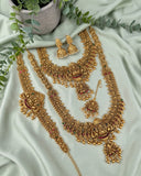 An ornate gold-toned Indian bridal jewelry set arranged on a light sage green satin fabric, featuring a necklace, bracelet, earrings, and a pendant with detailed filigree, gold beads, and small red and green stone accents. Decorative green leaves and a textured pot are placed around the set.