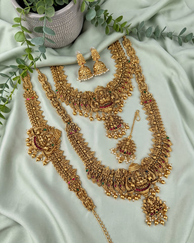 An ornate gold-toned Indian bridal jewelry set arranged on a light sage green satin fabric, featuring a necklace, bracelet, earrings, and a pendant with detailed filigree, gold beads, and small red and green stone accents. Decorative green leaves and a textured pot are placed around the set.