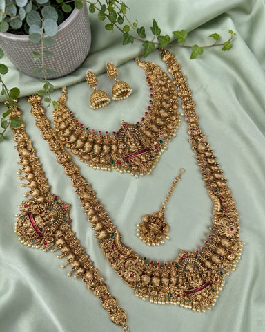 An ornate gold-toned traditional jewelry set arranged on pale green satin fabric, including a necklace, bracelet, earrings, and a matching pendant, with small red and green stone accents and pearl-like bead details. A potted plant is partially visible in the top left corner.