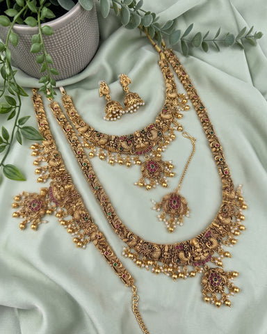 An ornate gold-toned traditional jewelry set arranged on pale mint-green satin fabric, with a necklace, matching earrings, bracelet, and pendant. The pieces feature intricate filigree, bead clusters, and small pink stone accents, with decorative green leaves and a textured pot in the background.