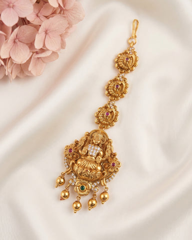 An ornate gold-toned traditional Indian jewelry piece, likely a maang tikka or pendant, decorated with a seated deity motif, small red gemstones, and green and white stones, displayed on a draped white fabric with pink flowers in the corner.