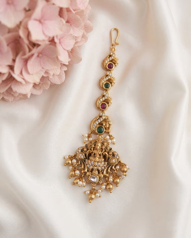 An ornate gold-toned Indian-style jhumka earring decorated with small green and red stones, white pearls, and a central clear gemstone, displayed on draped white satin fabric next to pink flowers.