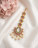 Gold-toned traditional Indian choker necklace with red and green stones and pearl-like drops, displayed on white satin fabric with pink flowers in the corner.