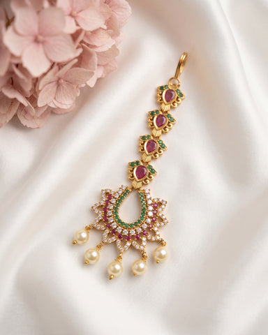 Gold-toned traditional Indian choker necklace with red and green stones and pearl-like drops, displayed on white satin fabric with pink flowers in the corner.