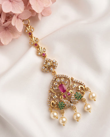 An ornate gold-toned traditional Indian choker necklace with a large teardrop-shaped pendant, decorated with clear crystals, pink and green stones, and dangling pearl-like beads, displayed on white satin fabric with pink flowers in the corner.